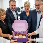 A group of five colleagues in a bright office setting gathered around a purple gift box filled with assorted cupcakes. The open box has a lavender lid printed with the text 'A little naughty, A lot of friends'.